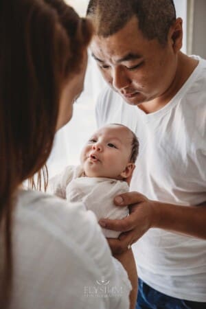 Parents hold their newborn baby boy between them as they stand at a bright window