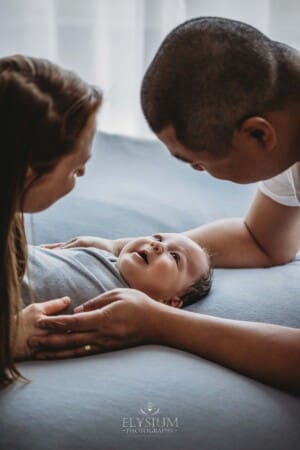 A newborn baby lays on a grey blanket smiling at his parents who are holding him