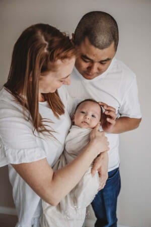 Parents hold their newborn baby boy wrapped in a white blanket and look adoringly at him