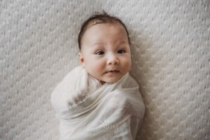 A newborn baby boy lays in a white wrap on a patterned white blanket
