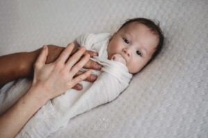 Parents hands hold their newborn baby boy as he lays in a white wrap on a textured white blanket