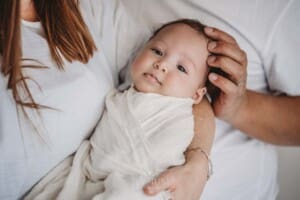 A newborn baby wrapped in white is held by his mum as his dad strokes his head