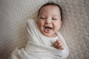A smiley newborn baby boy lays on a white textured blanket in a soft white wrap