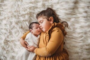 A little girl cuddles her baby sister as they lay on a fluffy white blanket