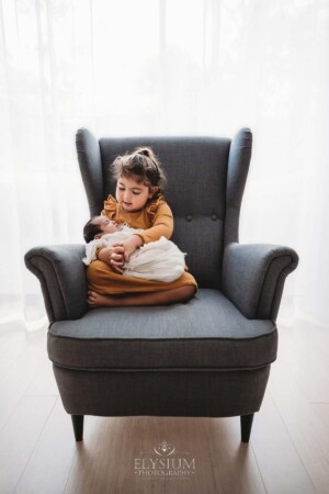 A child sits in a large grey armchair with her newborn baby sister in her lap wrapped in white