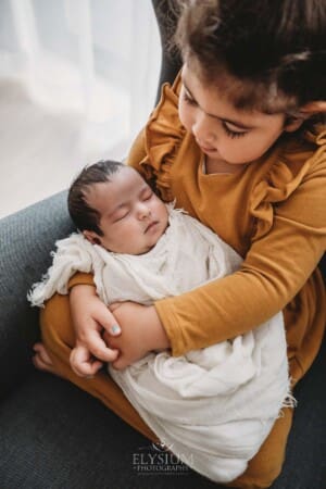 A child sits in a large grey armchair with her newborn baby sister in her lap wrapped in white