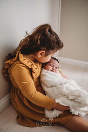 A child kisses her newborn baby sister on the forehead as they sit together on a white studio floor