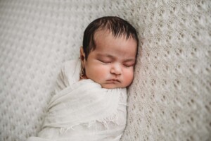 A newborn baby in a white wrap lays sleeping on a textured white blanket