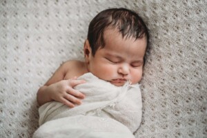 A newborn baby in a white wrap sleeping with one arm out on a textured white blanket