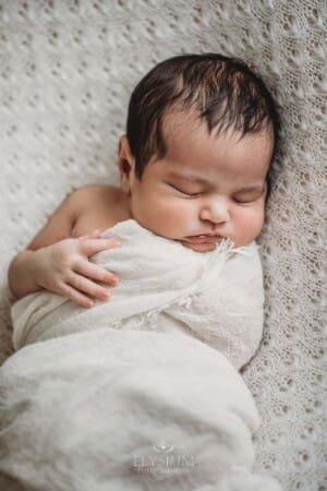 A newborn baby in a white wrap sleeping with one arm out on a textured white blanket