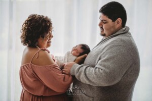 Parents hold their baby girl between them in their arms standing in front of a bright window