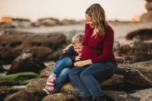 A pregnant woman sits on beach rocks and tickles her son as he giggles