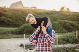 A little boy giggles as he sits on his dad's shoulders walking on a beach