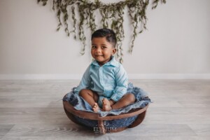 A baby boy sits in a wooden prop wearing blue with hanging leaves behind him