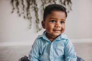 A baby boy wearing a blue outfit sits smiling in a studio with hanging leaves behind him