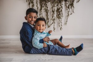 A boy holds his baby brother as they sit in a studio on the floor
