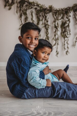 A baby boy is cuddled by his big brother as they sit in a studio with hanging green leaves behind them