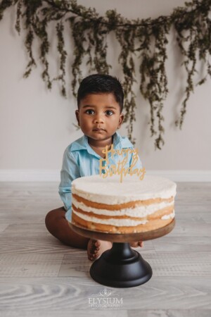 A baby boy waits to smash into his first birthday cake with hanging leaves behind him