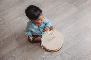 A baby boy plays with his birthday cake as he sits on a white studio floor