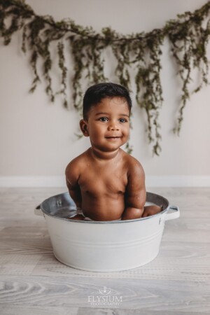 A baby smiles as he sits in a studio bath tub cleaning up after his cake smash