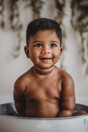 A baby sits in a white tub smiling as he splashes water around