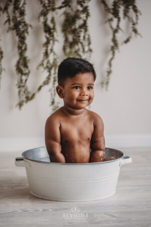 A baby boy has fun sitting in a white tub filled with bubbles after his cake smash