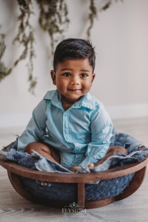 A baby boy sits in a wooden bowl on blue felt layers smiling in a studio