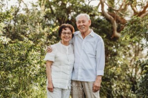 An elderly couple stand cuddling in a bushy park