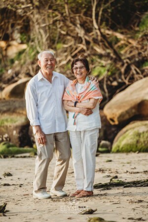 An elderly couple stand on a sandy beach at sunset