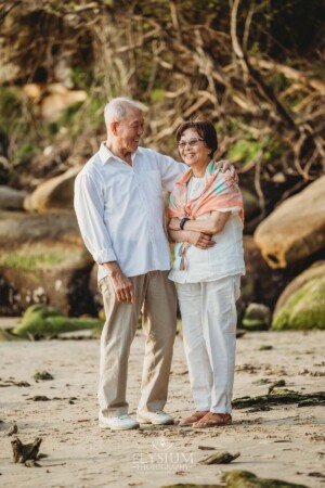 An elderly couple stand together laughing on a beach at sunset