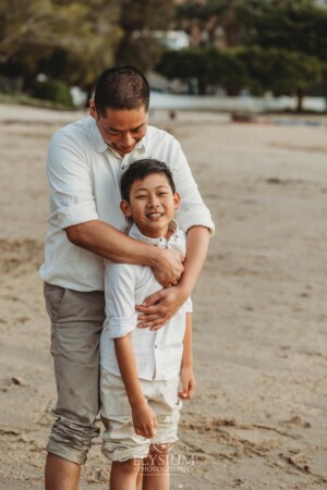 A father cuddles his son as they both stand on the shoreline at sunset