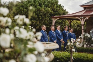 Groomsmen stand in front of rose bushes at a Camden wedding venue
