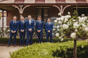 Groomsmen stand in front of rose bushes at a Camden wedding venue