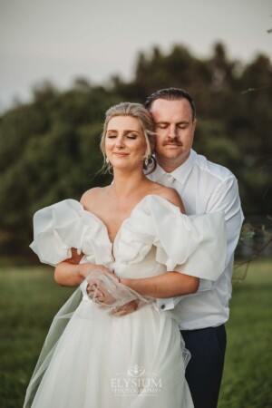 A groom hugs his bride from behind as they stand on a hill at sunset