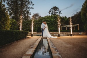 A bride and groom stand at the Burnham Grove fountain surrounded by fireworks