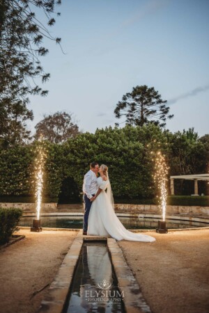 A bride and groom stand at the Burnham Grove fountain surrounded by fireworks