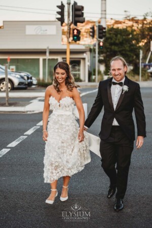 A bride and groom walk across a Sydney road towards their wedding reception at sunset