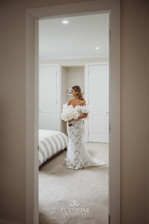 A bride stands framed by a doorway in her wedding gown holding a white bouquet