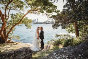 A wedding couple stand on the shore with views of Sydney Harbour behind them on a sunny day
