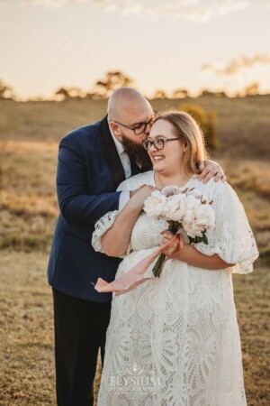 A groom kisses his bride on the cheek in a field at sunset