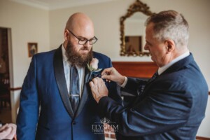 A groom is helped by his father to put on his jacket flower before the wedding