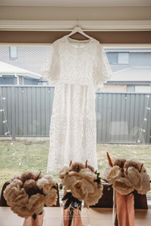 A lacey white wedding gown hangs in front of a bright window.