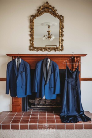 Navy blue wedding suits and a dress hang from a timber fireplace mantle