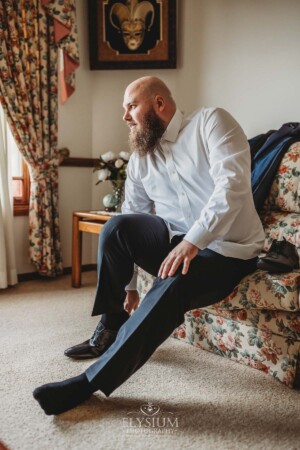 A groom sits on a floral sofa putting on shoes as he gets ready for his wedding day