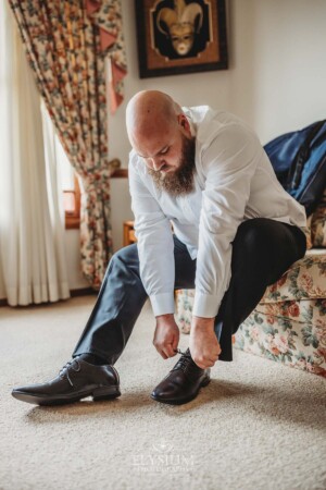 A groom sits on a floral sofa putting on shoes as he gets ready for his wedding day