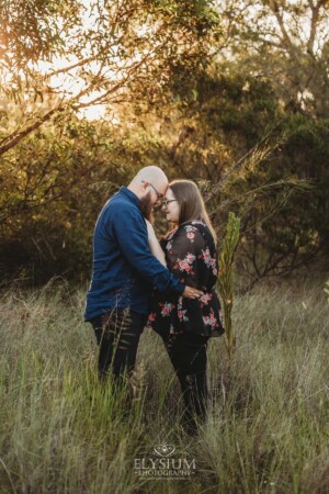 An engaged couple stand hugging in a long grassy field at sunset