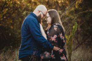 An engaged couple stand hugging in a long grassy field at sunset