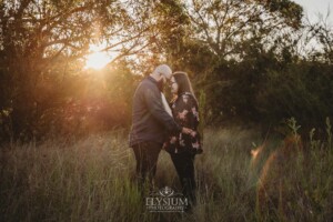 An engaged couple stand hugging in a long grassy field at sunset
