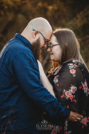 An engaged couple stand hugging in a long grassy field at sunset