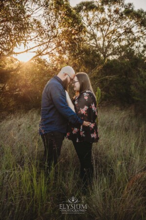 An engaged couple stand hugging in a long grassy field at sunset